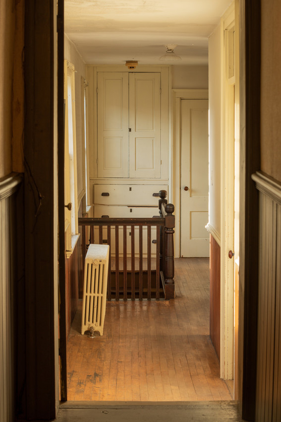 Hallway through a doorway, with a radiator left, stairwell railing, and behind, a white-painted wood built-in cabinet over three drawers, next to a door. 