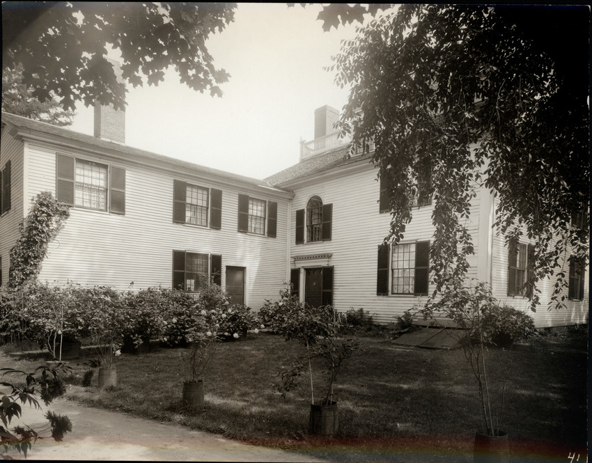 The rear of a two-story white building with dark shutters, showing the service wing perpendicular to the main house with a square lawn in between.