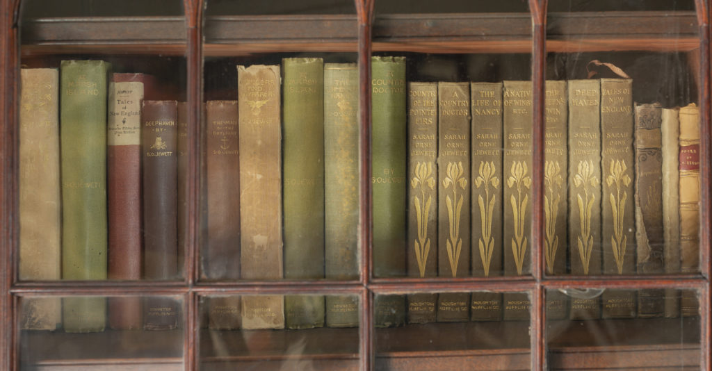 Old hardcover books of varied colors, several with gold designs on the spins, in a wooden case with a glass front