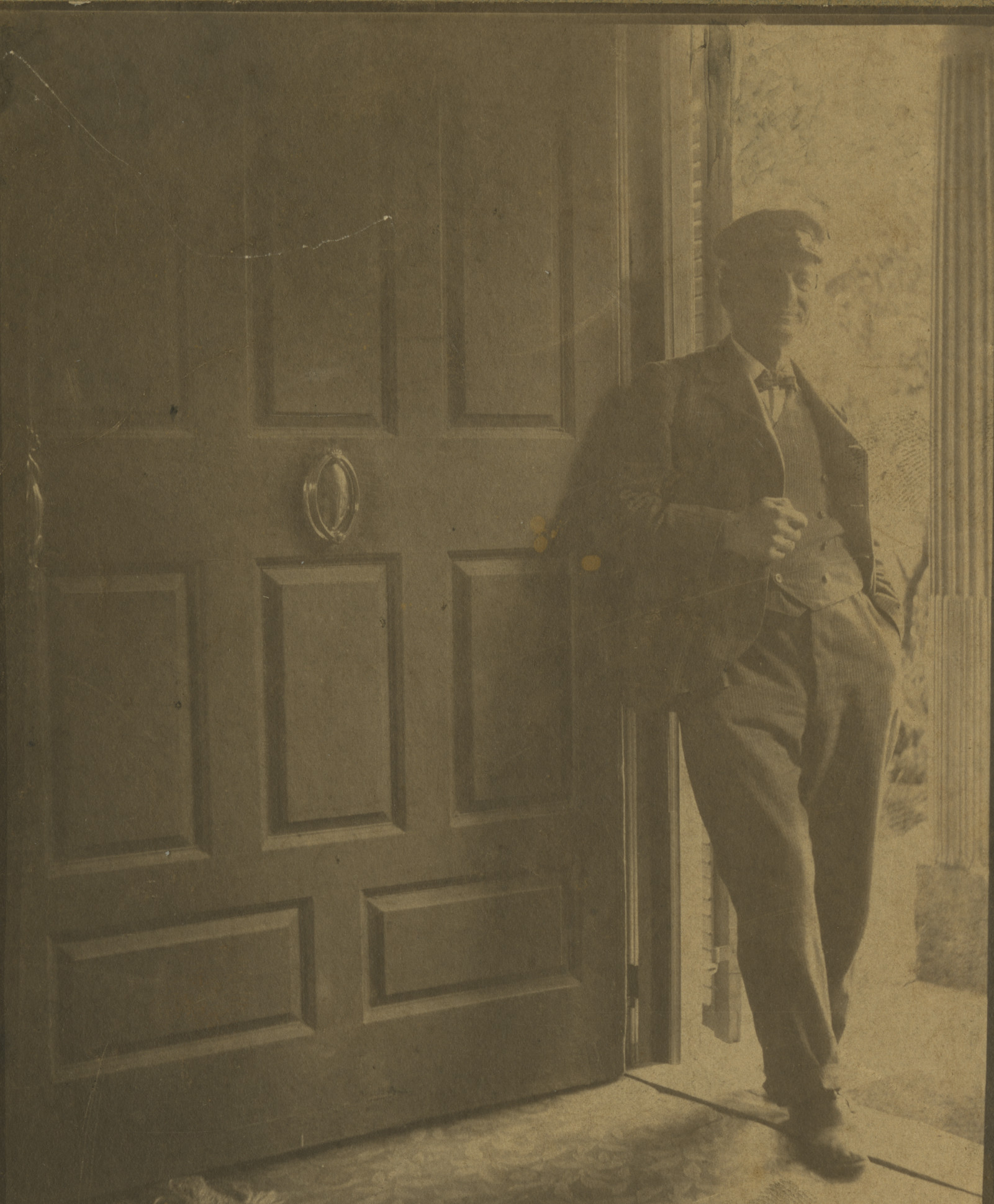 A man wearing a suit with vest, bow tie, and brimmed cap, leaning against a large, open door with a door knocker.
