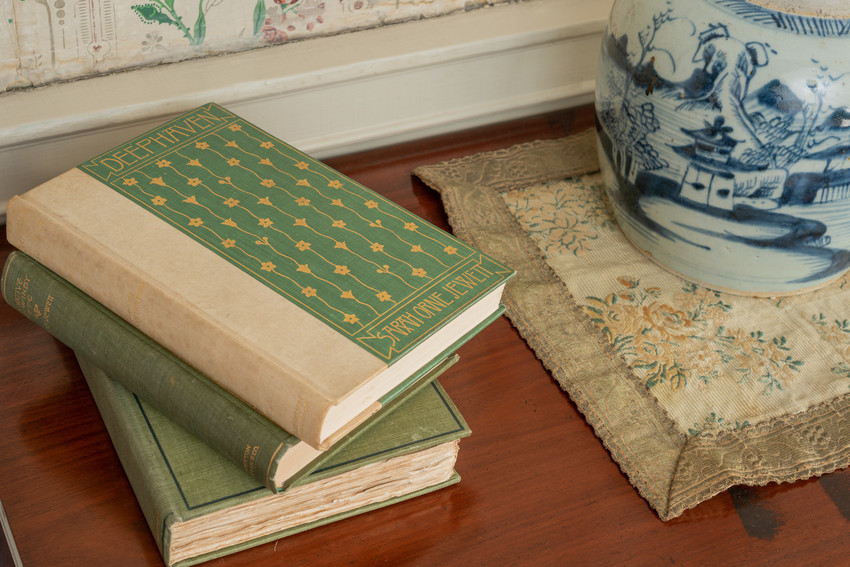 A  stack of three books, the top of which is a green volume with a gold floral design, Deephaven by Sarah Orne Jewett,  next to a blue and white decorative vase on a wooden table. 