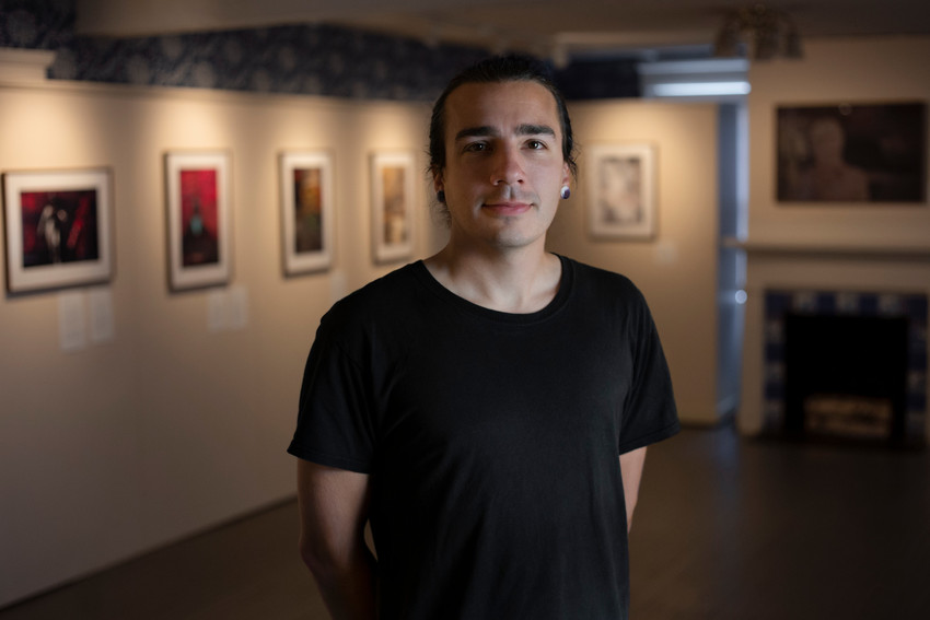 Photograph of artist Lakotah Sanborn. Half-length e of man in black shirt, with black hair standing in a gallery of art.